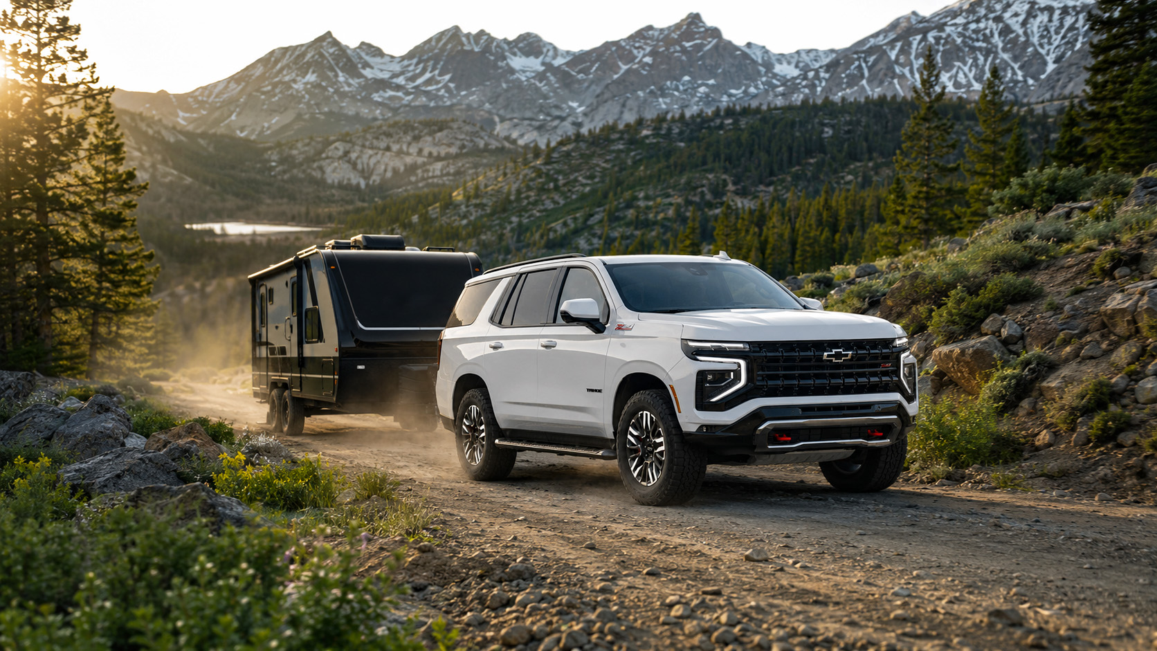 A white 2026 Chevrolet Tahoe Z71 towing a black travel trailer on a mountain road at sunrise, with snow‑capped peaks and evergreen trees in the background, illustrating Tahoe’s power and off‑road performance.